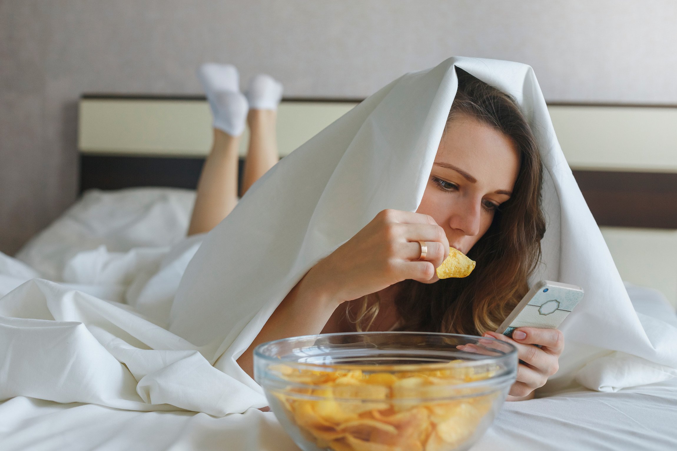 A woman lying in bed on her stomach looking at her phone and eating potato chips while under the covers