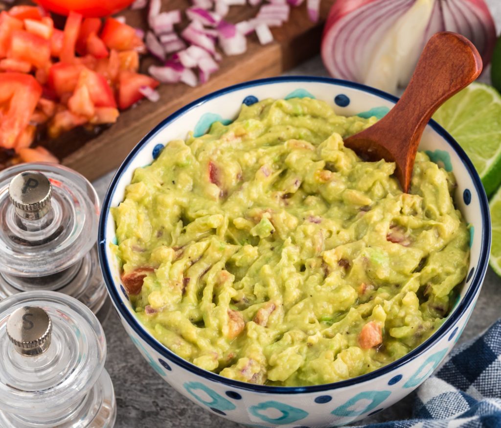 FRNs Homemade Guacamole plated in a ceramic dish near salsa, seasonings, and diced onions
