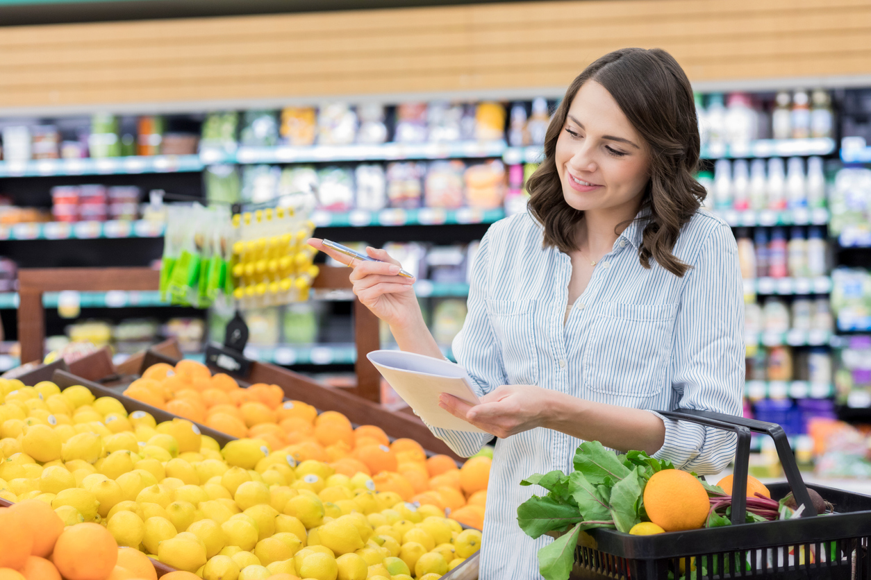 A cheerful young woman holds a pen and shopping list as she stands in the produce section of a grocery store. She has a shopping basket on her arm as she checks her list.