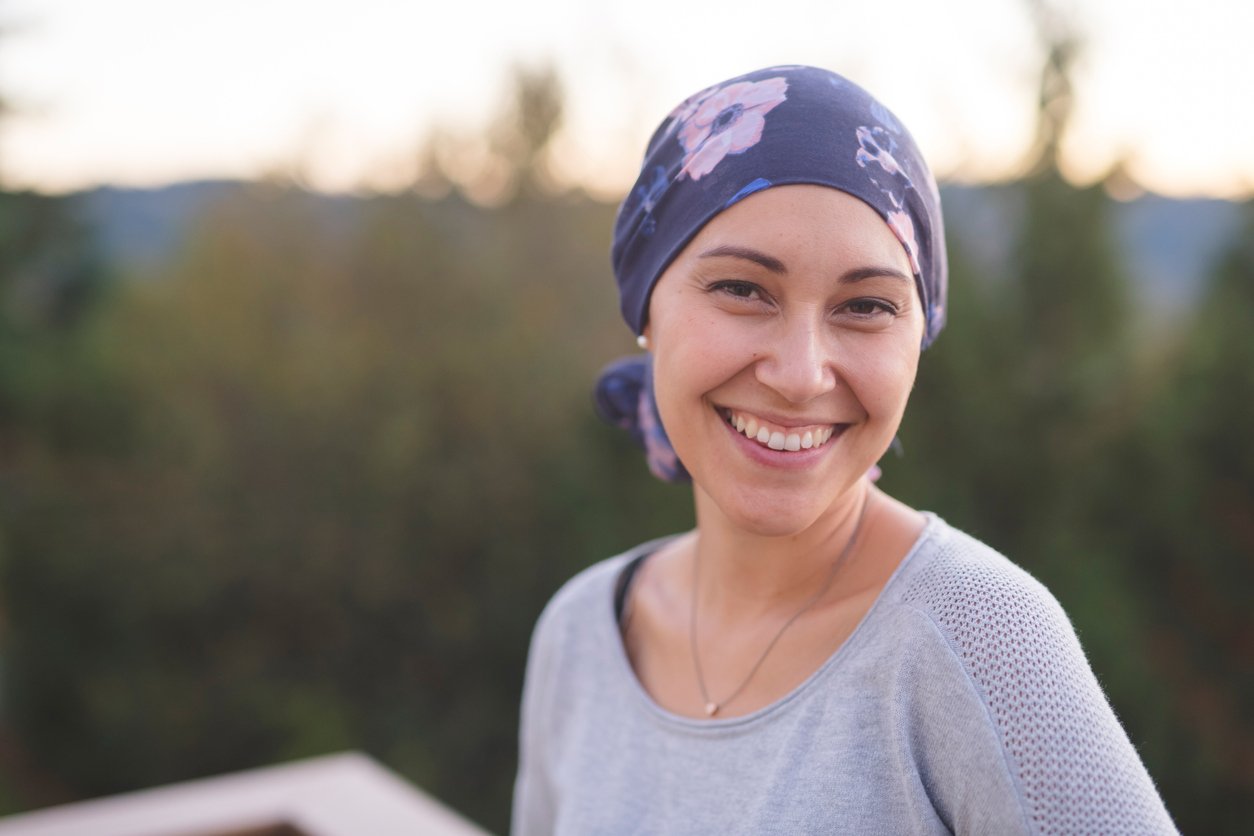 A beautiful young woman wearing a head wrap looks toward the camera and smiles radiantly. She is standing outdoors and there are mountains and trees in the background.