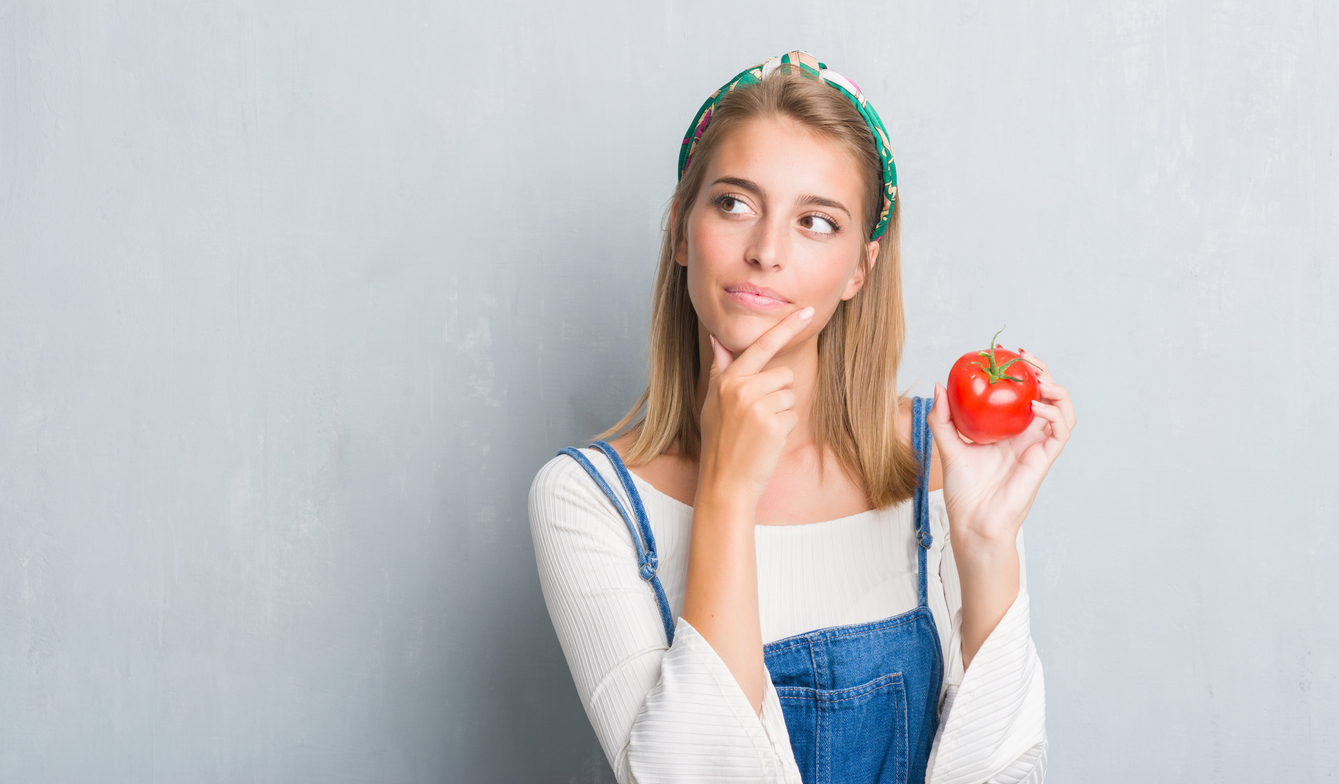 Beautiful young woman over grunge grey wall eating fresh tomato serious face thinking about question, very confused idea