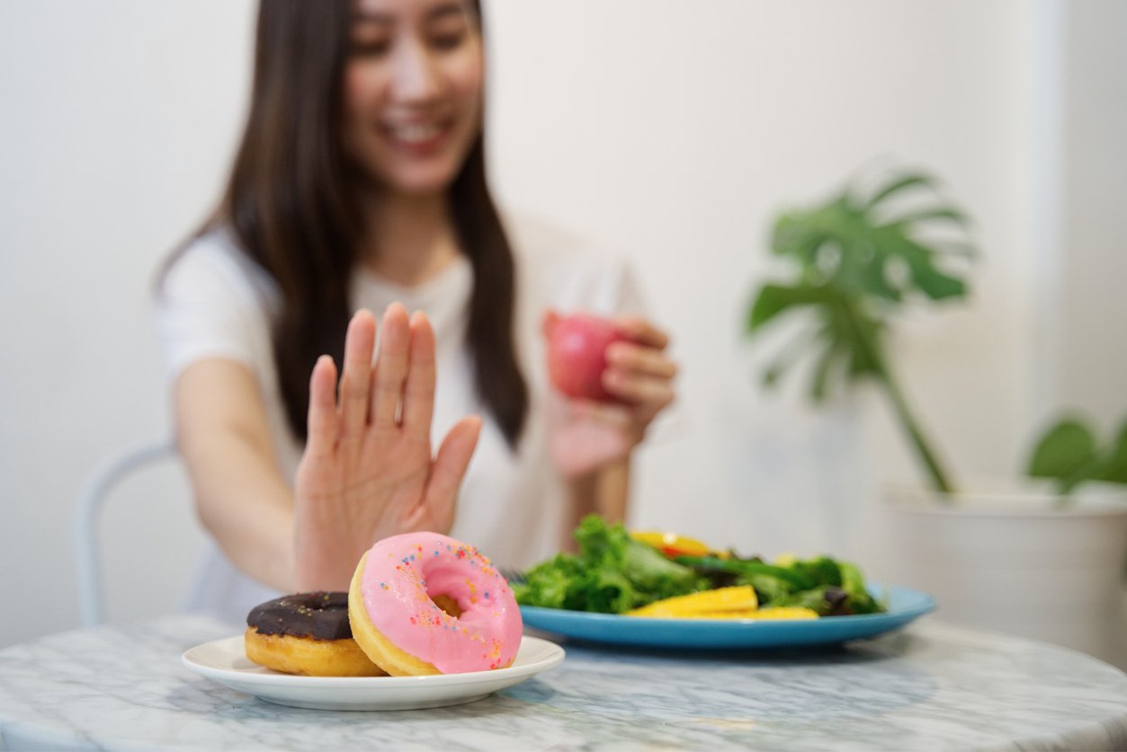 年輕女孩為了健康而節食的概念。特寫女性用手推開她最喜歡的甜甜圈拒絕垃圾食品，選擇紅蘋果和沙拉以獲得良好的健康。