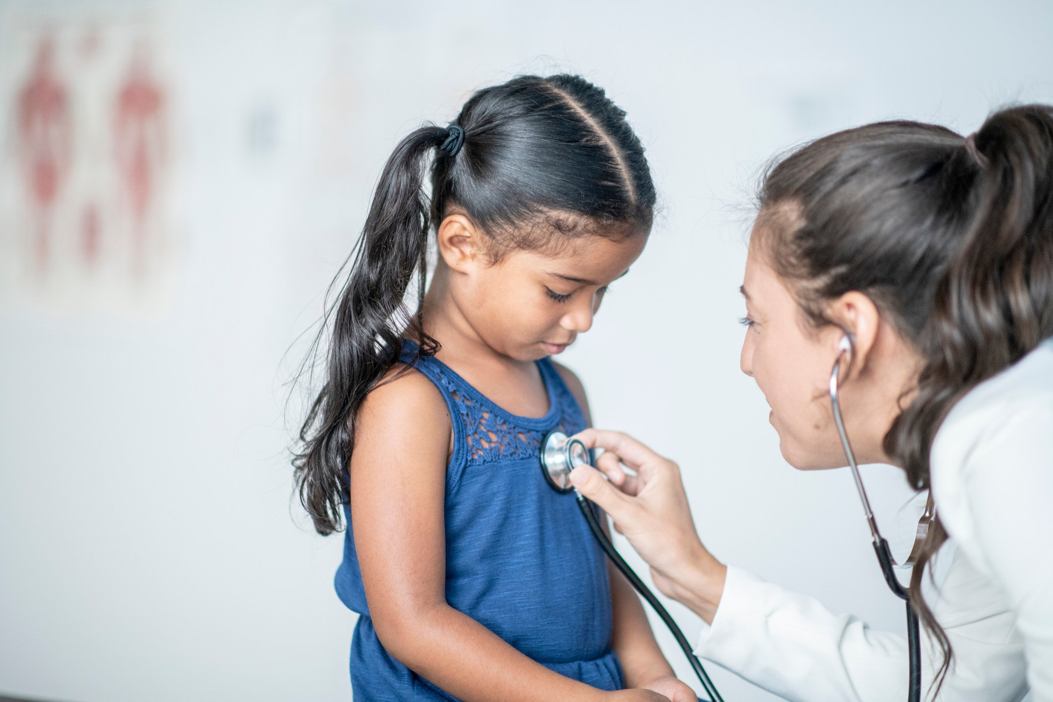 A young hispanic girl looks down at the stethoscope that her doctor is placing on her chest. Her doctor is a young caucasian woman and is wearing professional clothing.