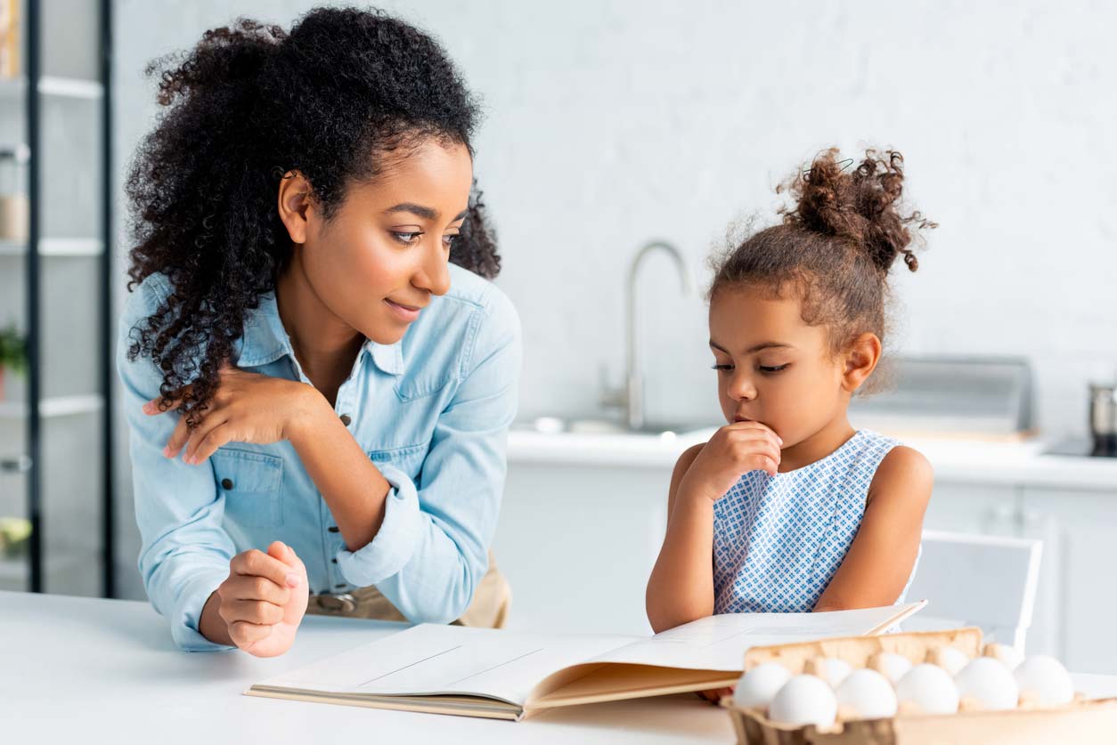 Mother and daughter looking at cookbook together with eggs on counter.