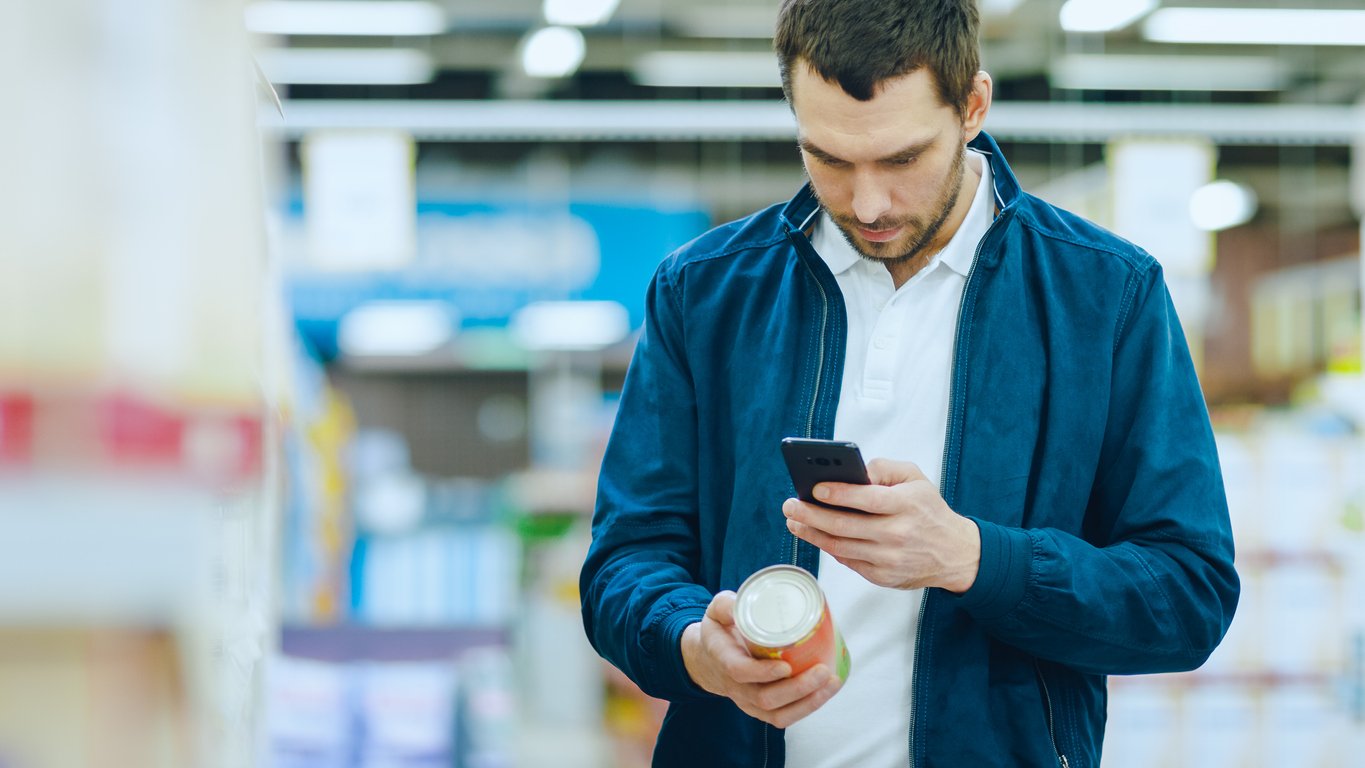 At the Supermarket: Handsome Man Uses Smartphone to Check Nutritional Value of the Canned Goods and Buy it. He's Standing with Shopping Cart in Canned Goods Section.