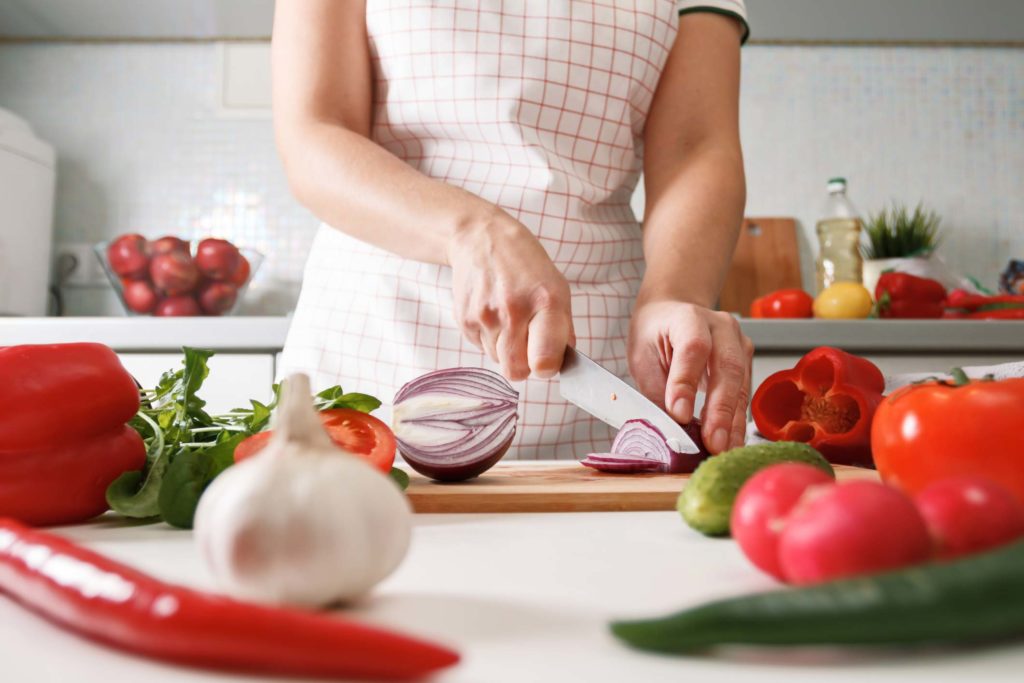 Woman slicing onion on cutting board in the kitchen, surrounded by vegetables