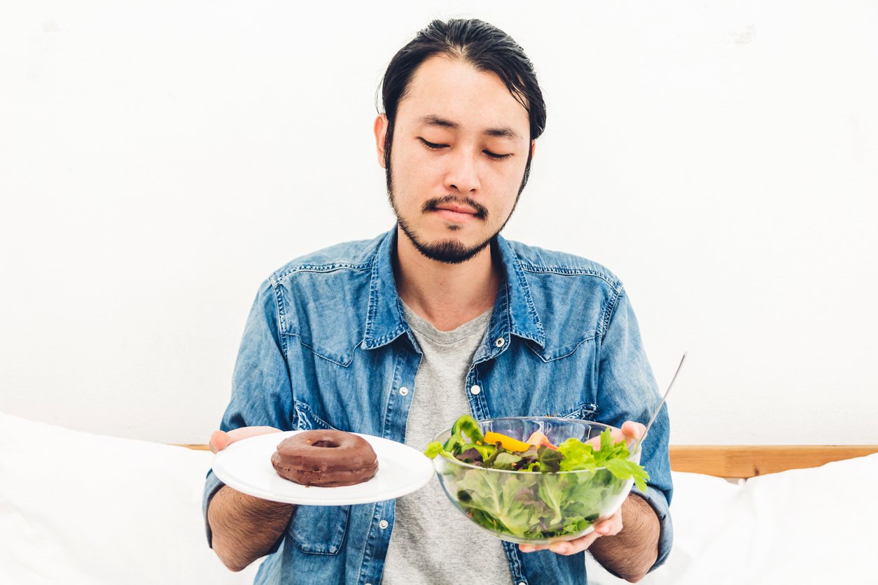 Man holding and making choice between healthy salad and calorie bomb chocolate donut on bed at home.Healthy eating and Junk food concept