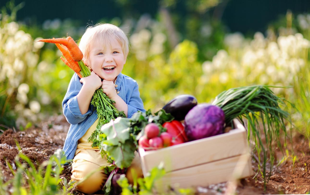 happy child holding carrot next to box of vegetables