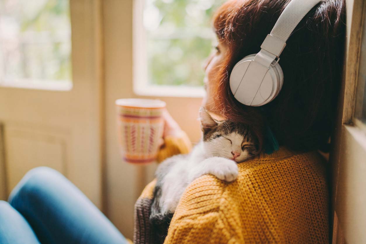 Woman wearing headphones, snuggling a cat, and drinking a hot drink