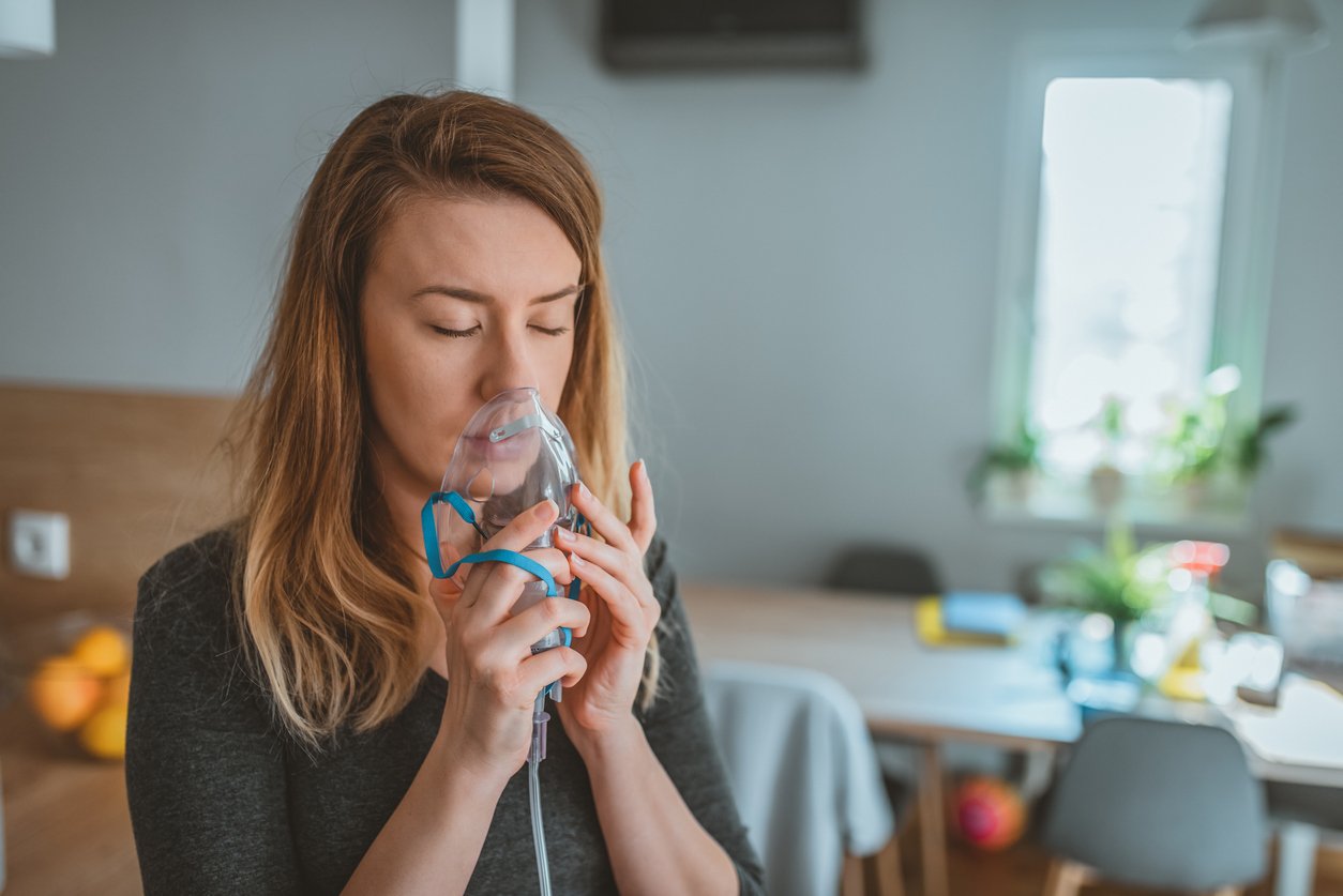 Photo of woman makes inhalation at home, holding a mask inhaling fumes spray the medication into lungs sick patient. Self-treatment of the respiratory tract using inhalation. Young woman's face inhaling through inhaler mask. Brown hair female patient inhalation therapy by the mask of inhaler with soft stream smoke from asthma inhaler.