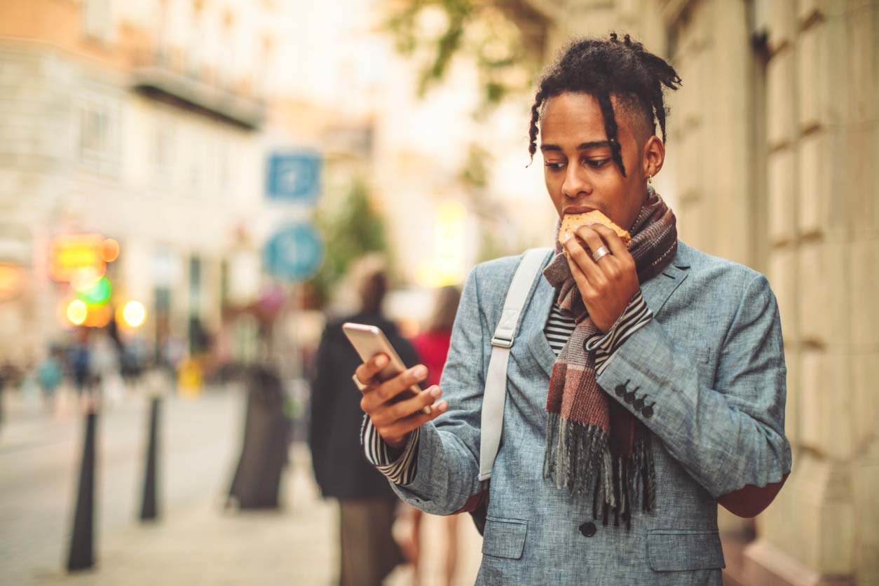 Person of color eating sandwich while holding phone on sidewalk