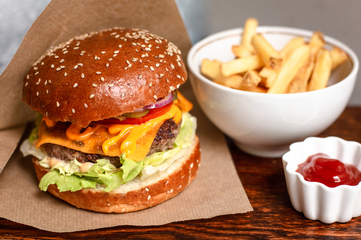 Homemade beef burgers with cheese, tomato, red onion, pickled cucumber and lettuce served on wooden board with french fries and ketchup. Close up.