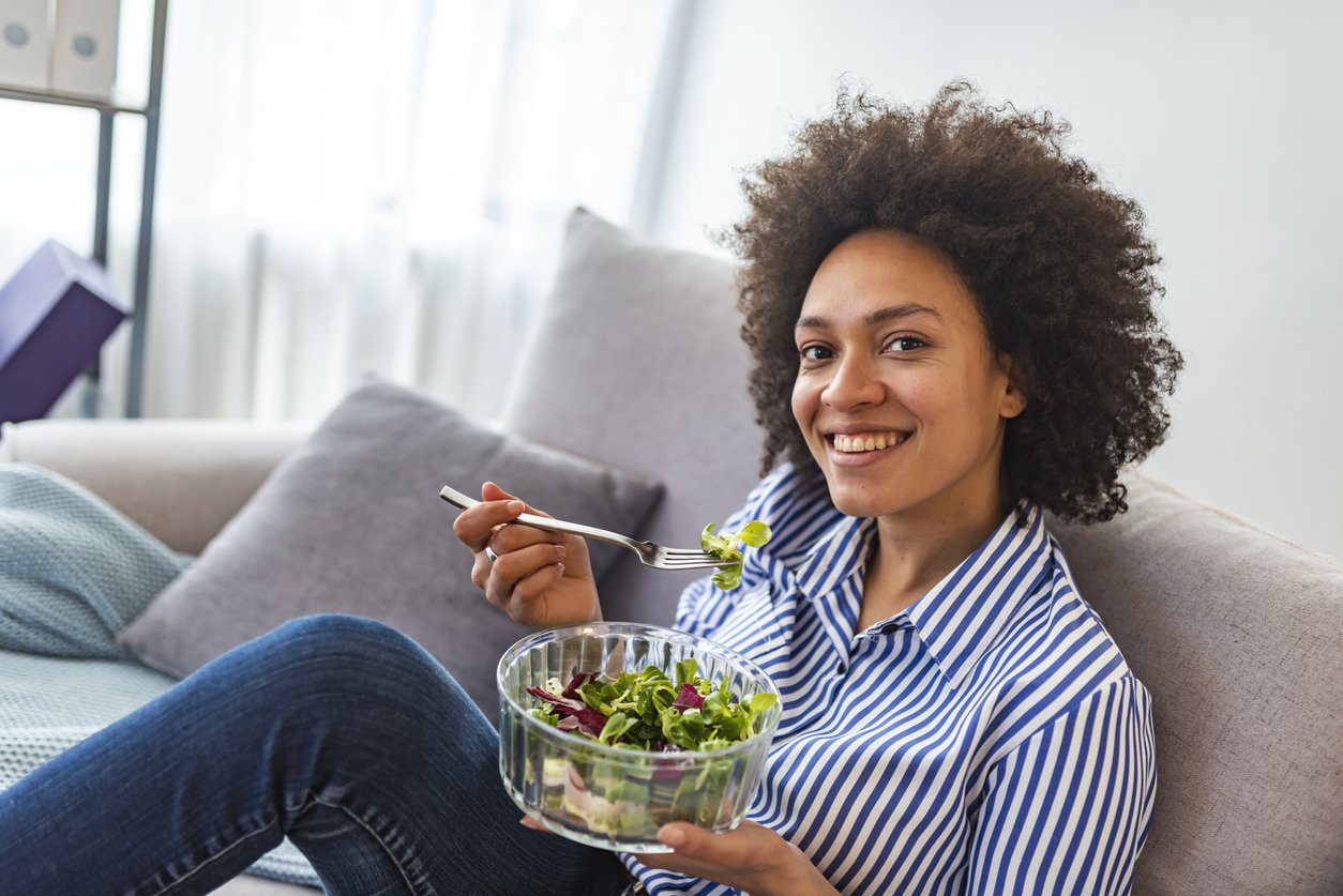 Cheerful young Afro american woman eating vegetable salad at home. Close-up Of Beautiful African American Woman Eating Salad At Home. Beautiful woman on the sofa eating a healthy salad