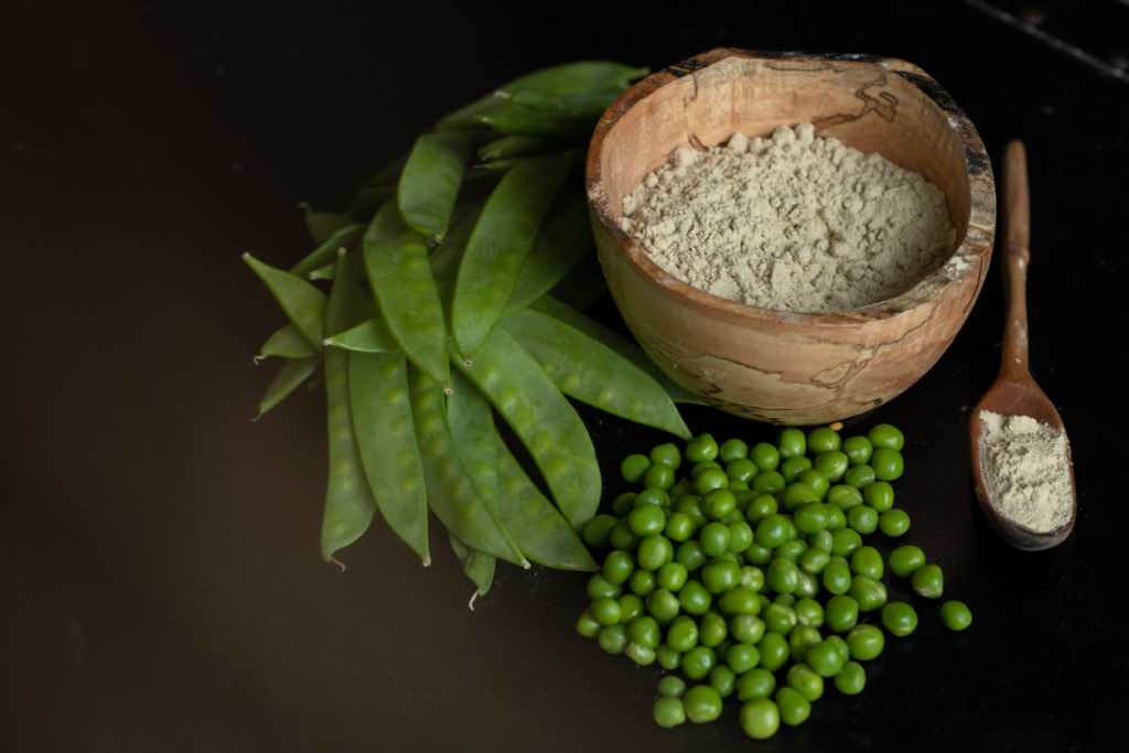 Pea protein powder in a wooden bowl with a pile of snow peas and green peas