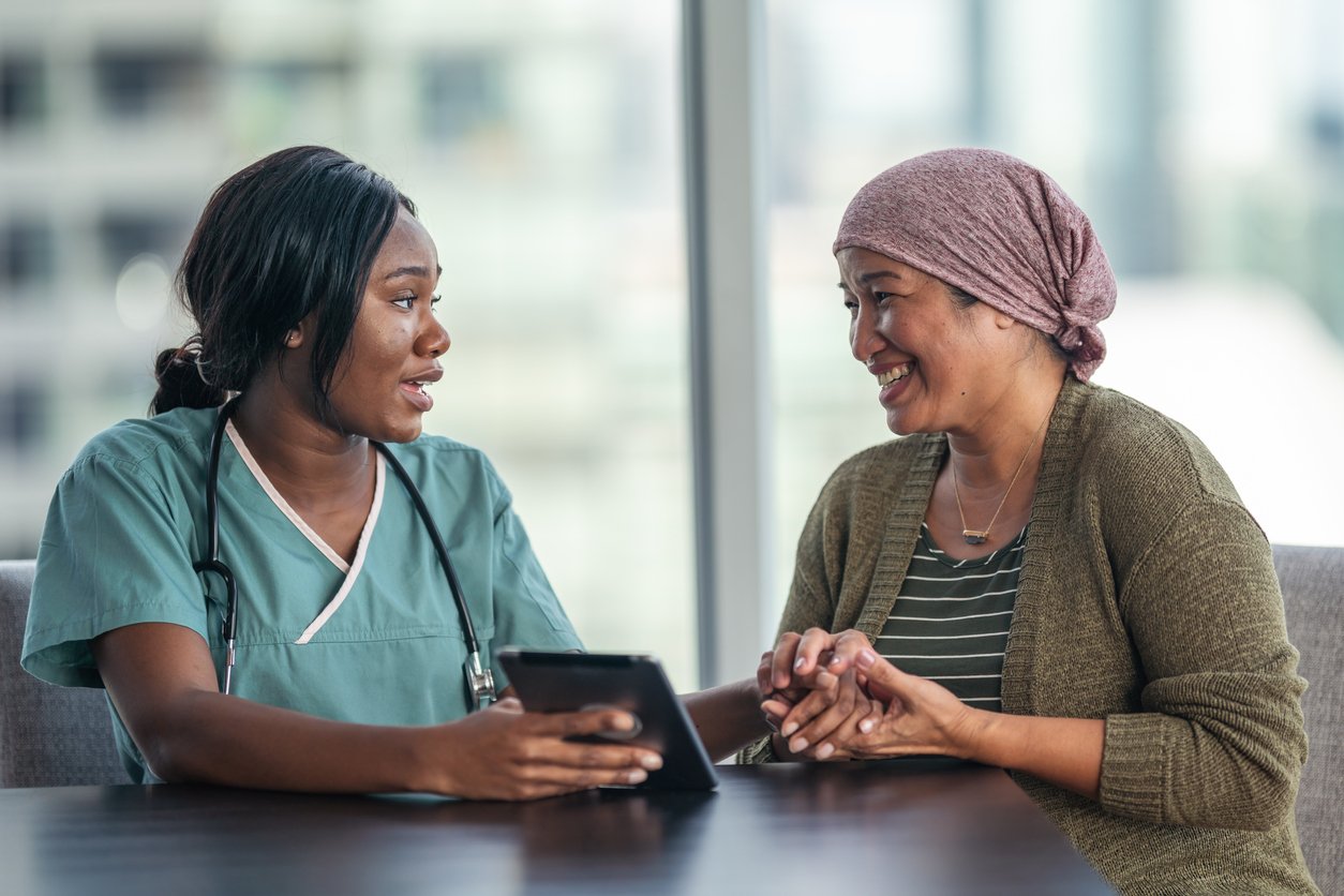 An Asian woman with cancer is consulting her doctor. The two women are seated at a table together. The patient is wearing a bandana to hide her hair loss. The medical professional is showing the patient test results on a digital tablet. They are discussing a treatment plan.