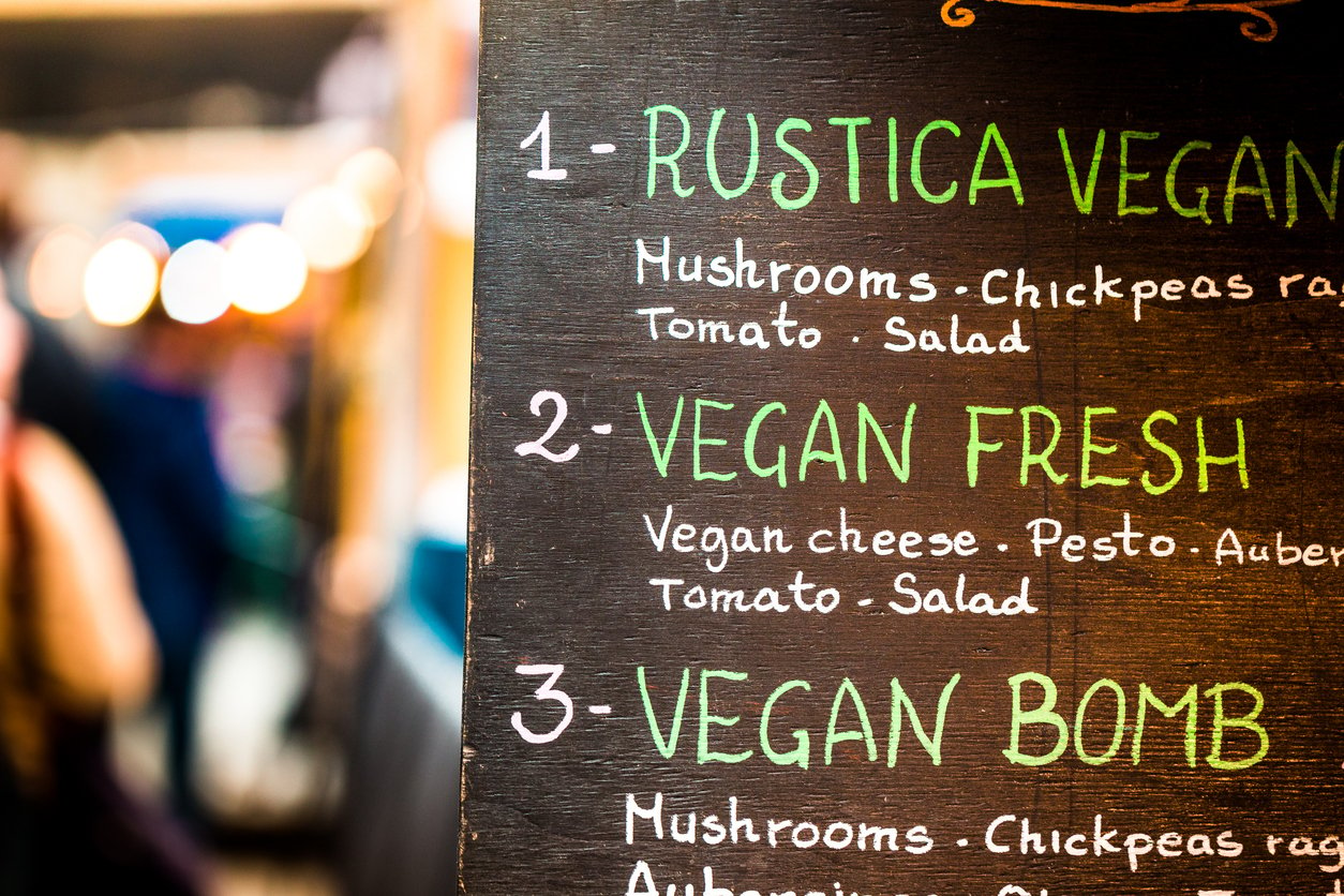 Close up image depicting menu options for a vegan and vegetarian cafe on a city street in central London, UK. Menu options include such things as vegan cheese, mushrooms, aubergines and olives. Focus is on the sign with colorful chalk writing in the foreground, while the street is defocused beyond. Room for copy space.