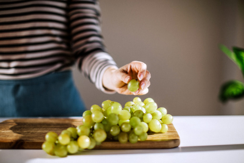 Woman holding green grape over cutting board full of grapes in the kitchen