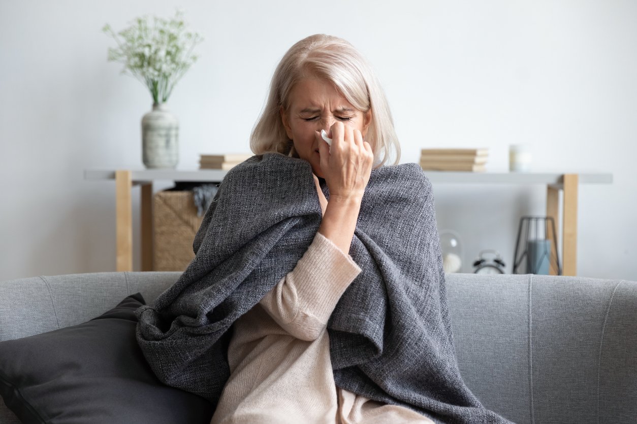 Middle-aged 50s sick frozen woman seated on sofa in living room covered with warm plaid sneezing holding paper napkin blow out runny nose feels unhealthy, seasonal cold, weakened immune system concept