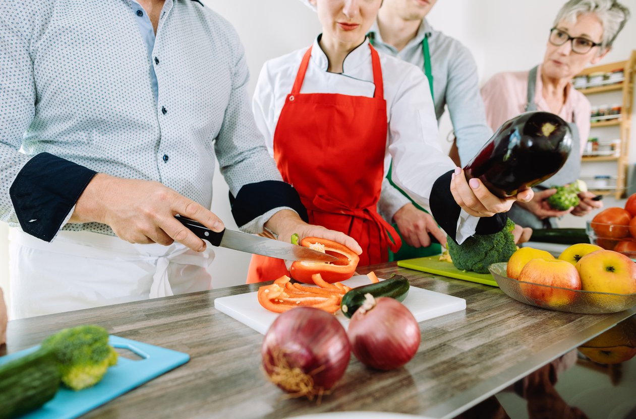 Man in training kitchen cutting vegetables under watchful eye of dietician