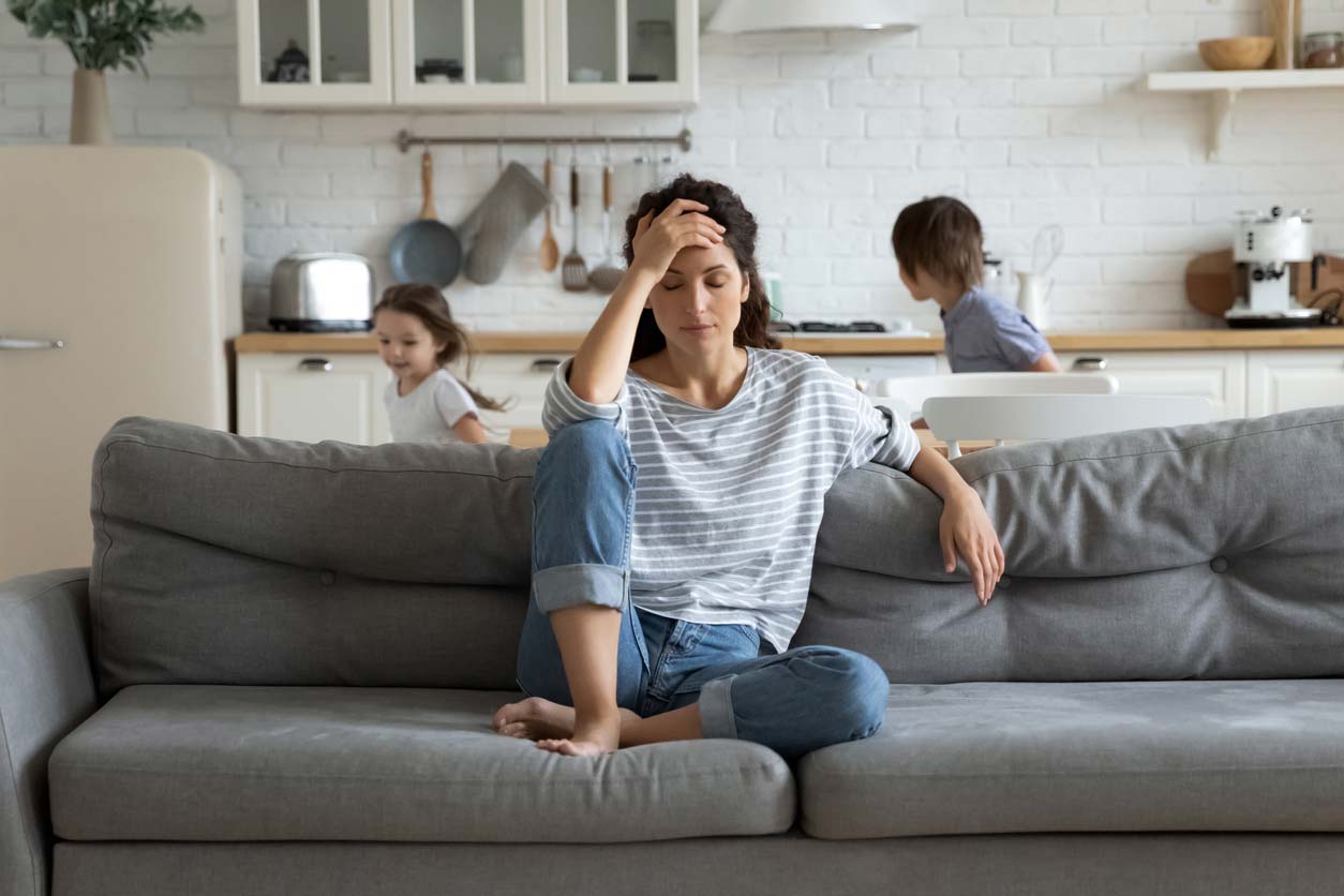 Exhausted mother sitting on couch while kids running in background - stress can impact hormones