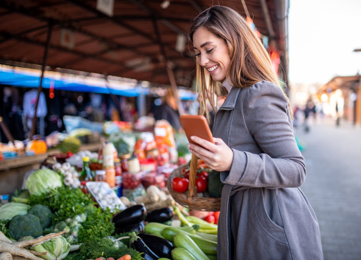 в руке и в другом смартфоне со списком продуктов, стоя на фермерском рынке.