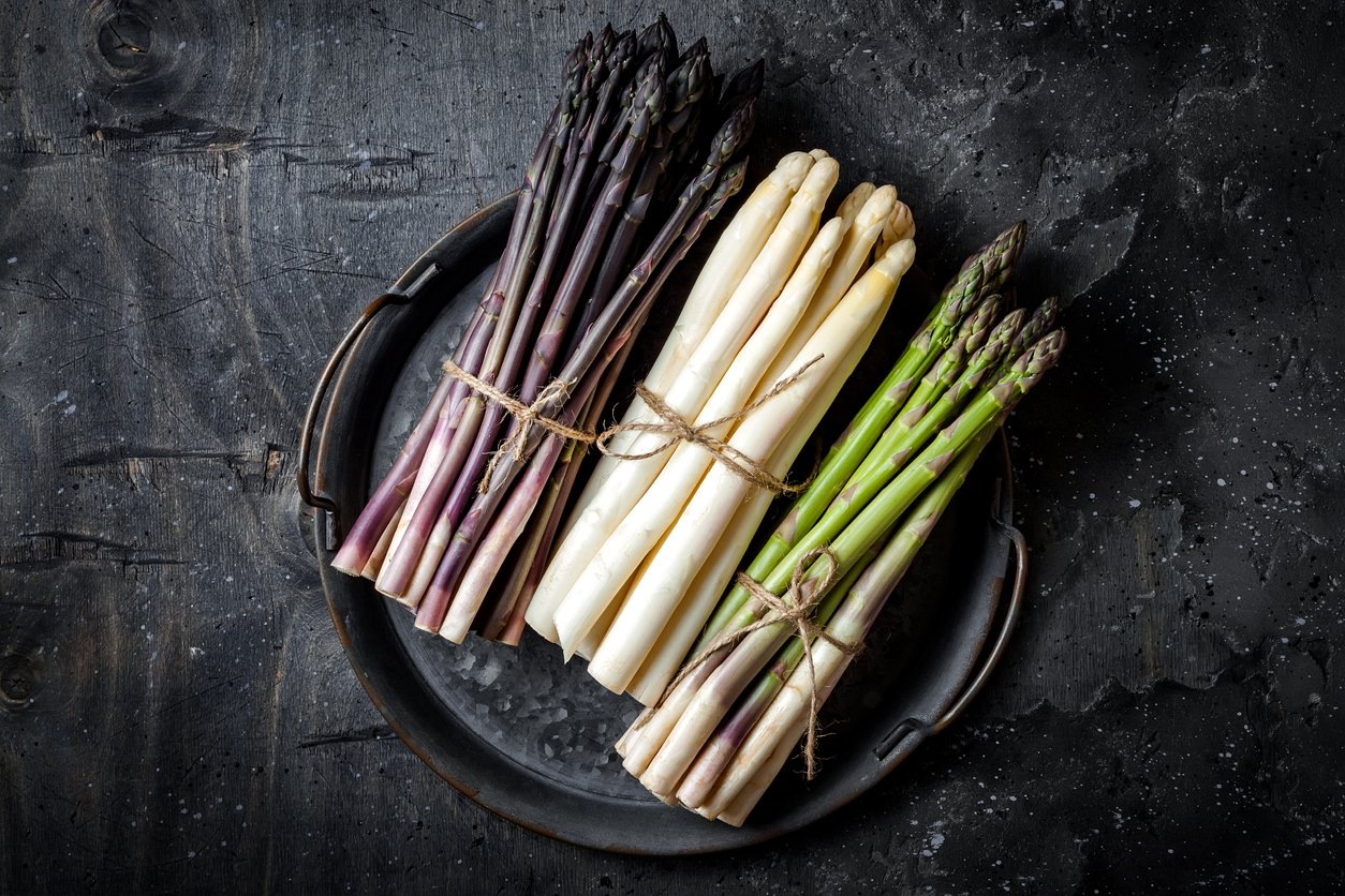 Bunches of fresh green, purple, white asparagus on vintage metal tray over dark grey rustic background. Top view, copy space