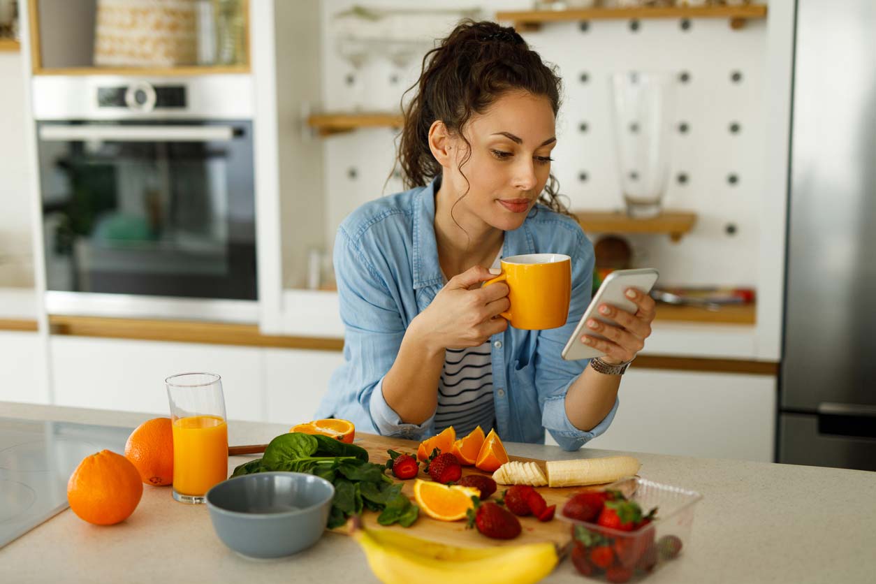 woman using phone and drinking tea while preparing greens and fruit