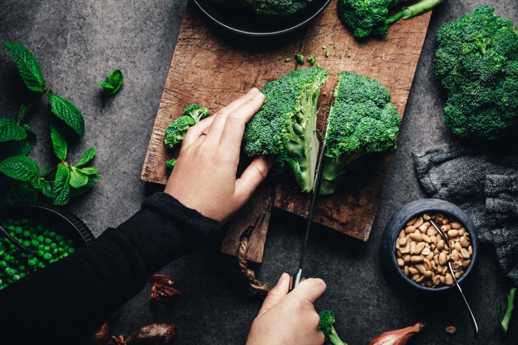 Hands cutting broccoli with knife on a cutting board, surrounded by folate-rich food