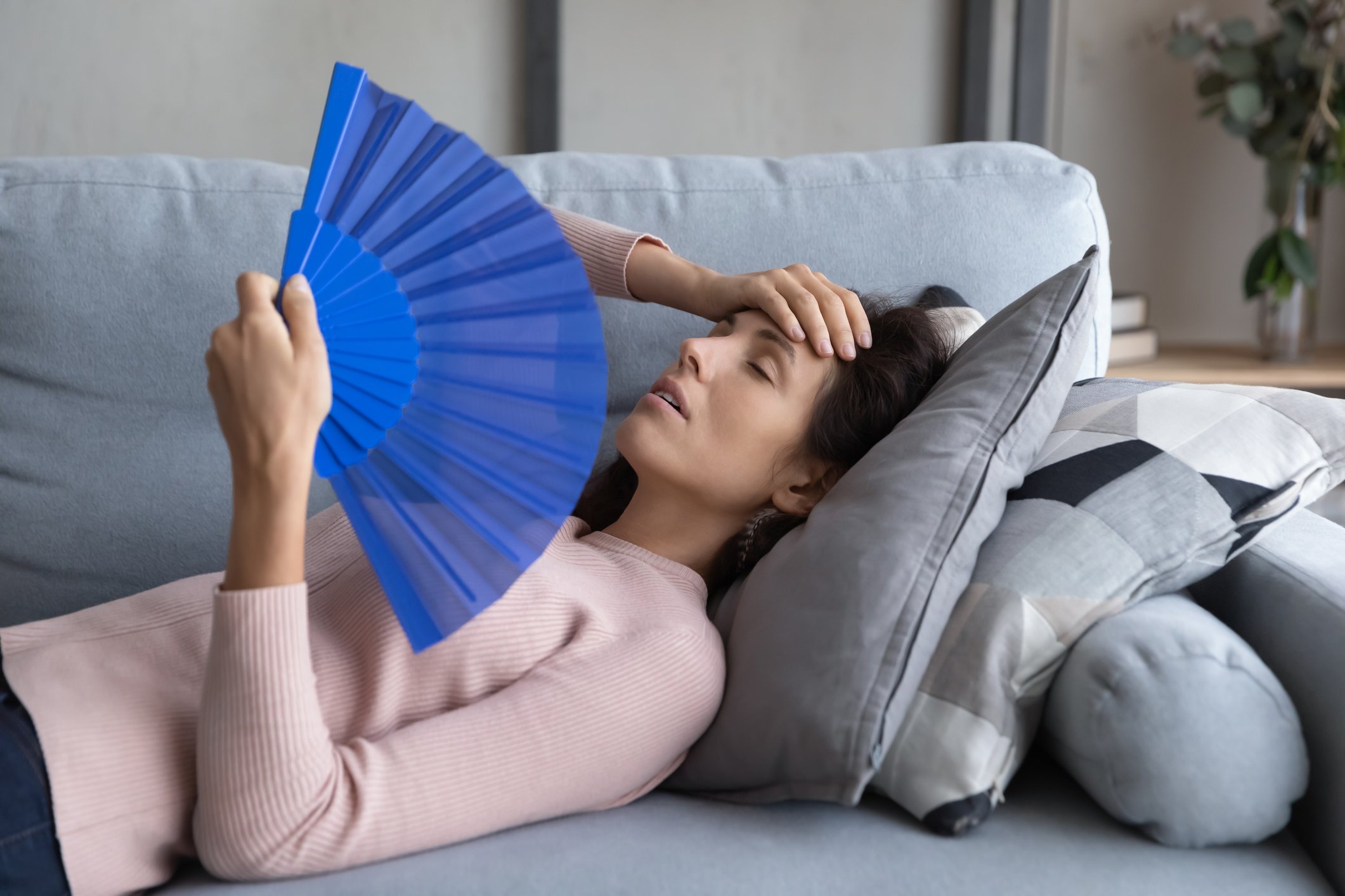 Unwell tired young woman lying on sofa at home wave with hand fan, suffer from hot weather, lack of air conditioner, exhausted overheated girl use waver, have hormonal imbalance, health problems