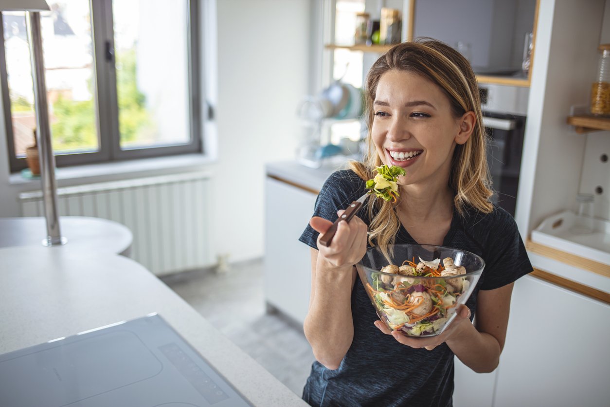 Photo of cheerful woman standing with salad dish at home kitchen. Beautiful girl eating fresh salad at modern apartment. Closeup young woman enjoying vegetarian food indoors.