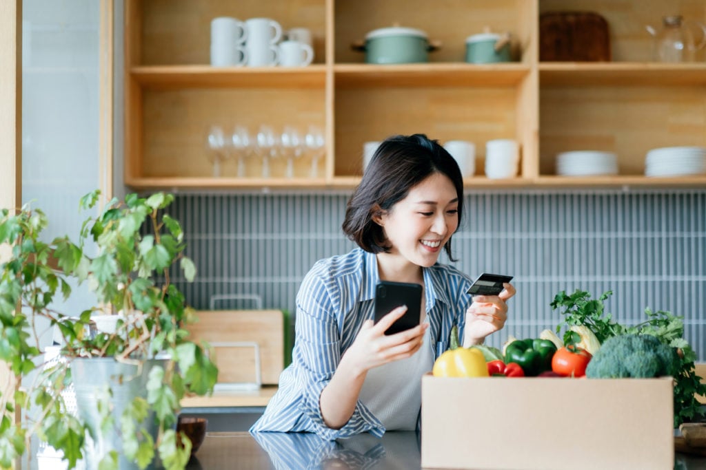 Asian woman shopping online and holding her credit card in the kitchen