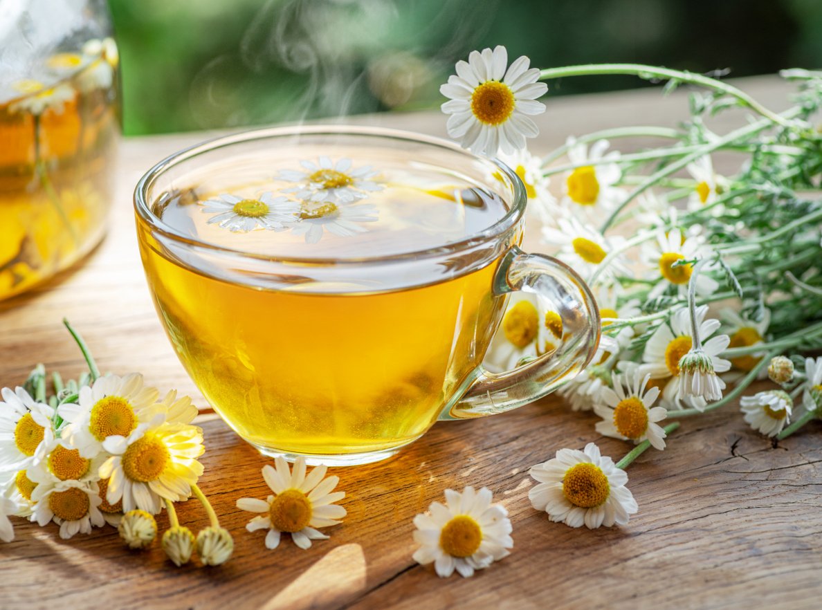 Herbal chamomile tea and chamomile flowers near teapot and tea glass on wooden table. Countryside background.