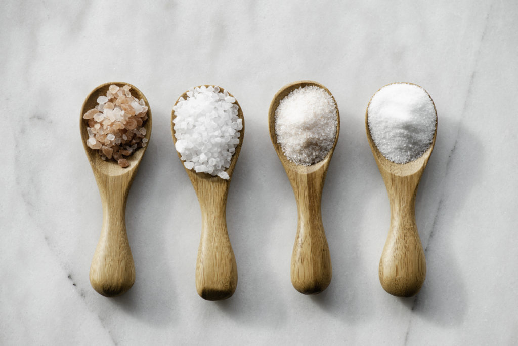 Various salt types in 4 wooden spoons on white marble.