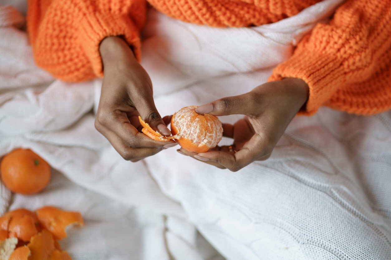 afro woman hands peeling ripe sweet tangerine wear orange sweater lying in bed under