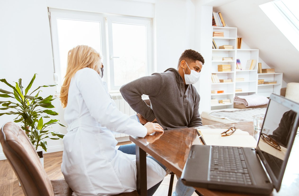 Young African-American man being examined by female doctor in a doctor's office. Man with protective face mask complained of kidney pain.