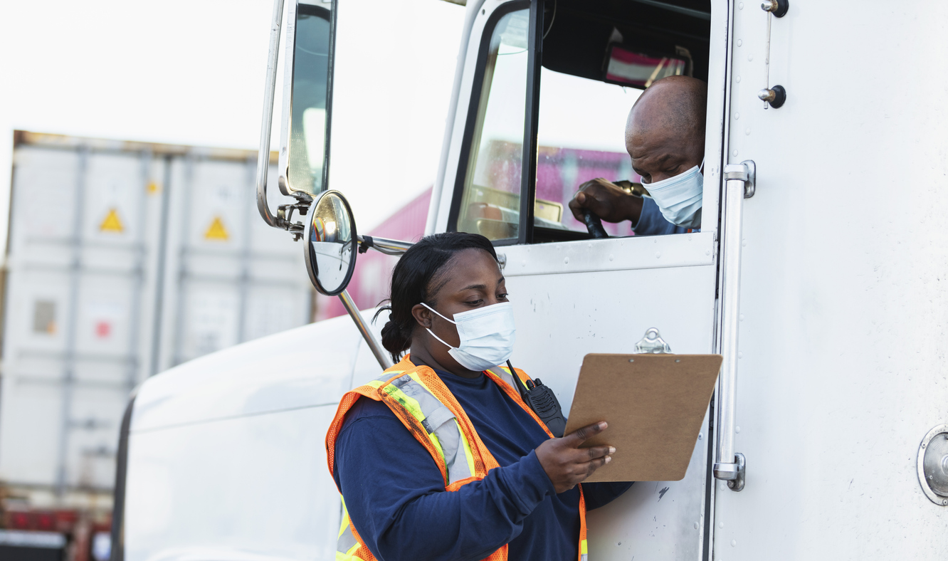 Two African-American workers in their 40s at a shipping port conversing. One is a truck driver, leaning out the open window of his semi-truck. He is talking to a woman standing next to the truck, a dock worker or manager coordinating deliveries. They are looking at the clipboard she is holding. They are wearing protective face masks, working during COVID-19, trying to prevent the spread of coronavirus.