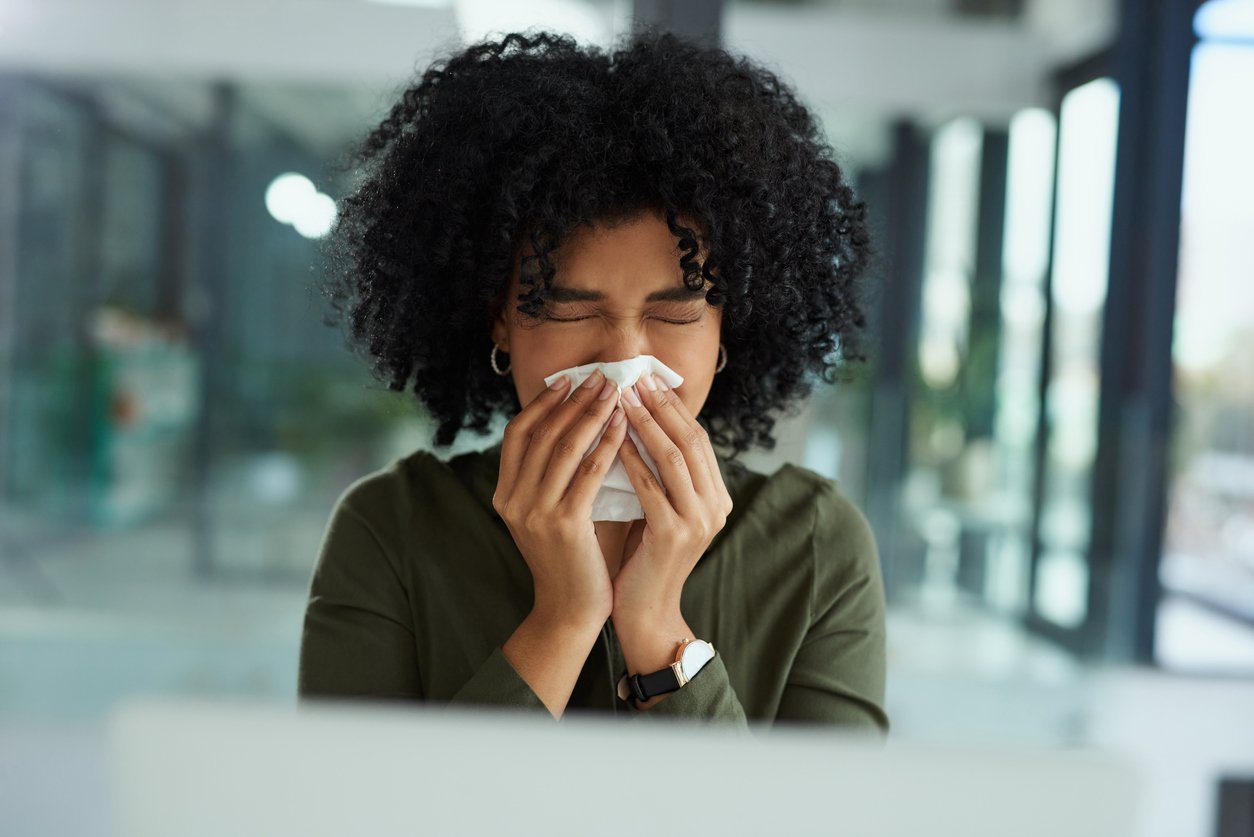 Shot of a young businesswoman blowing her nose while using a laptop in a modern office