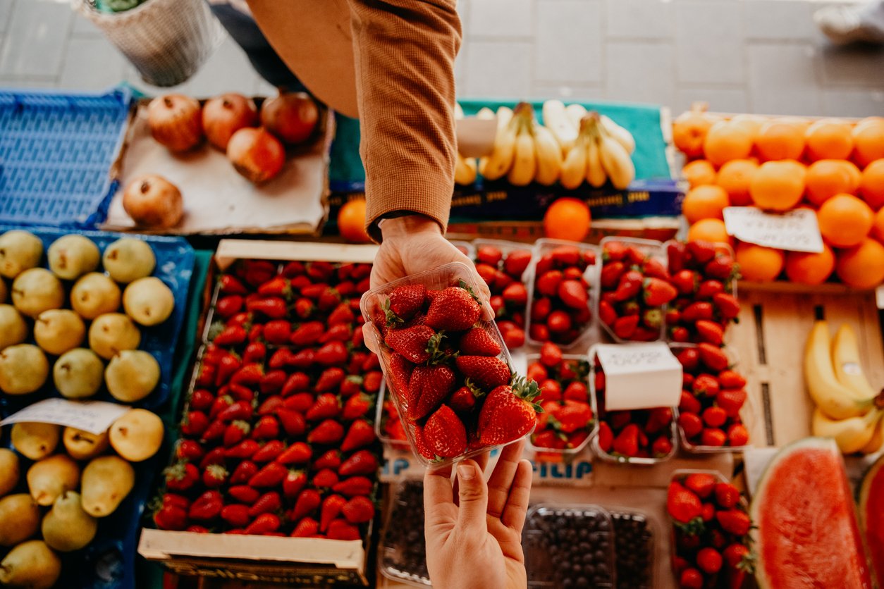 An app view photo of a male hand giving A box of fresh strawberries ￼￼to a salesman hand, while the rest of the fresh fruits is looking great on the market table ￼
