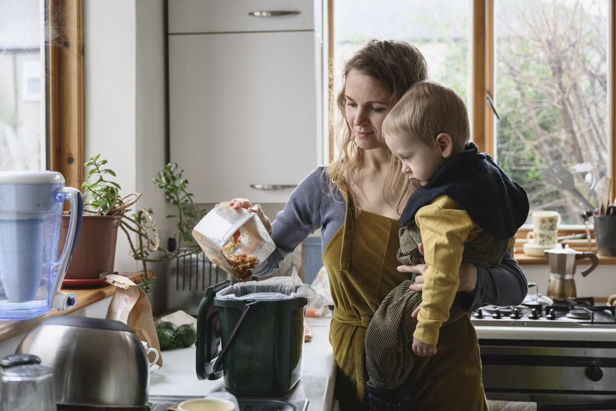 Waist-up view of mid 30s Caucasian woman standing in domestic kitchen holding toddler in her arms and adding unused food to compost bucket.