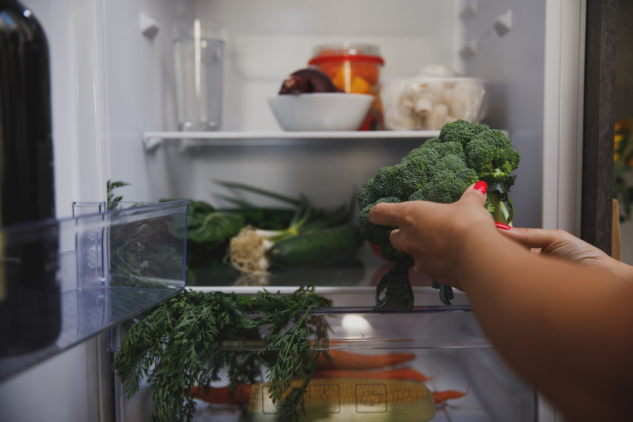 Close up shot of unrecognizable woman putting a head of broccoli on the shelf in her fridge while unloading groceries.