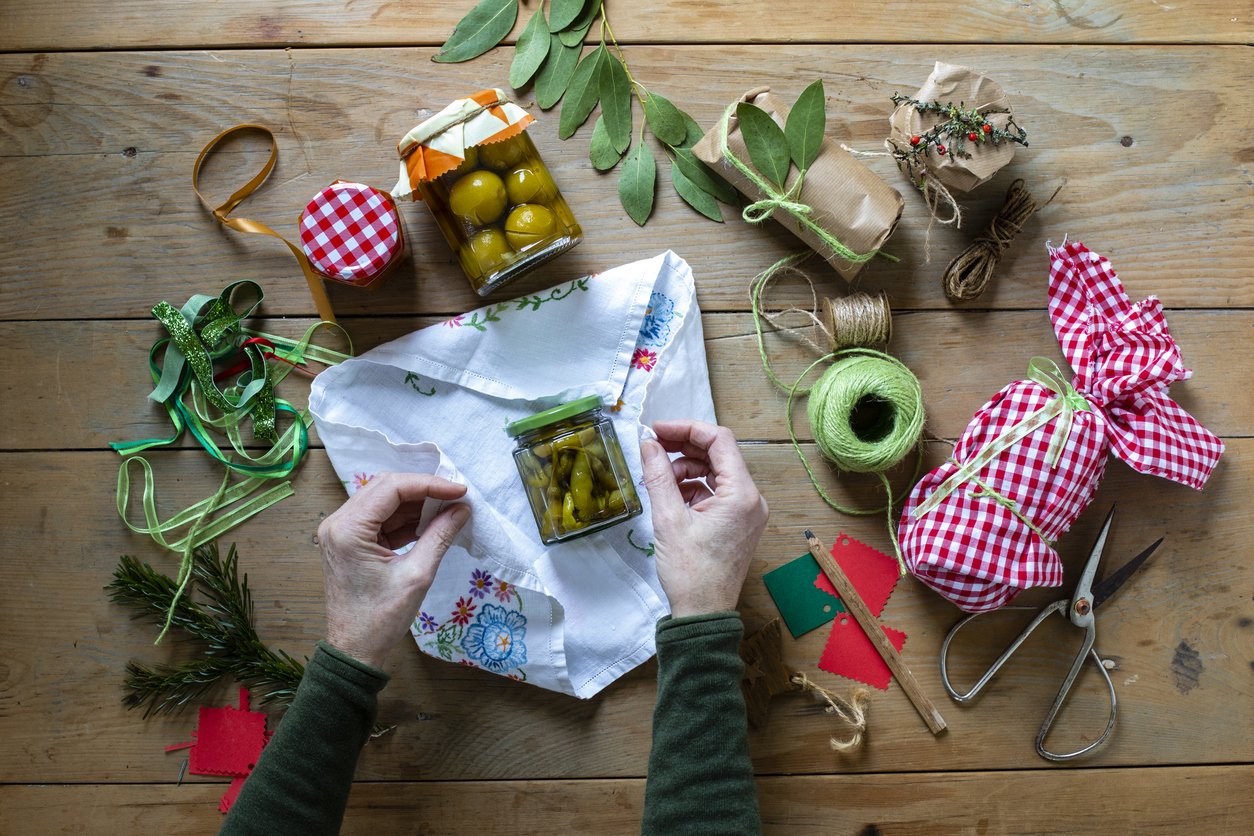 A table top view of an unrecognisable woman wrapping a jar of homegrown chilli peppers in embroidered material on a wooden table top, she is using string, ribbon, eucalyptus leaves and holly to decorate the gift. Other presents on the table are wrapped and others are unwrapped.