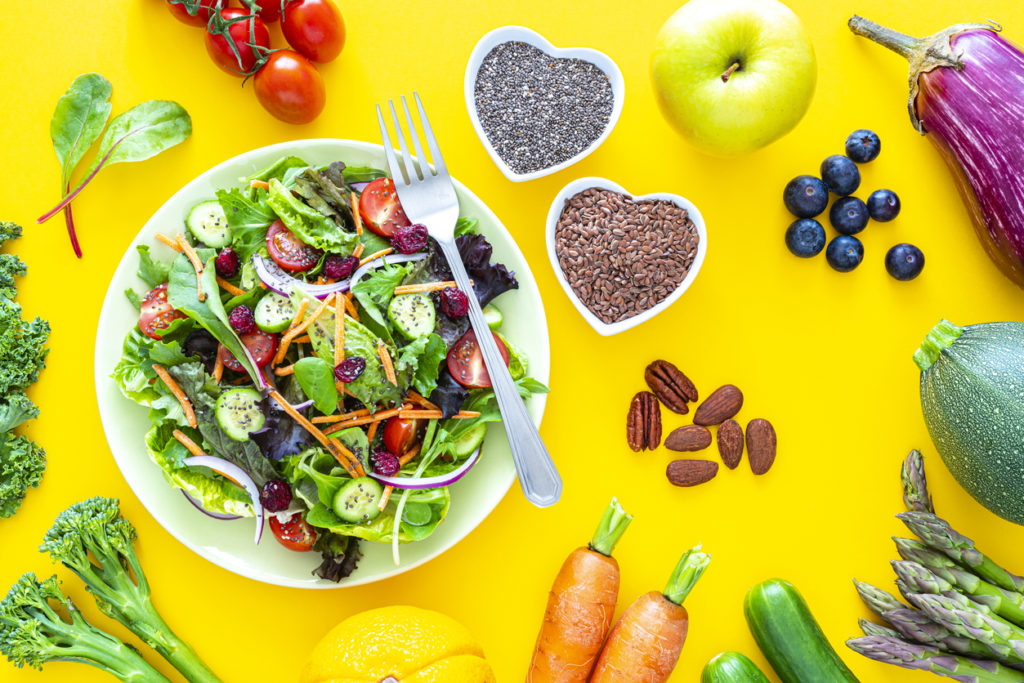 Overhead view of a fresh salad plate shot on yellow background with fruits, vegetables and seeds are scattered on the table.