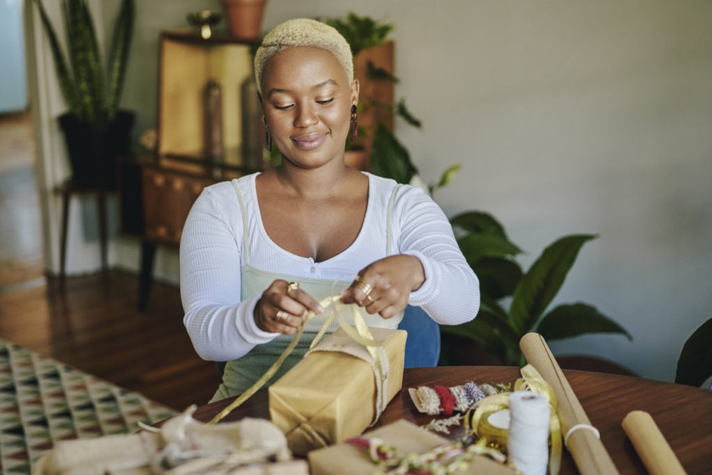 Smiling young woman sitting at a table at home and using recycled paper and ribbons to wrap Christmas presents for the holidays