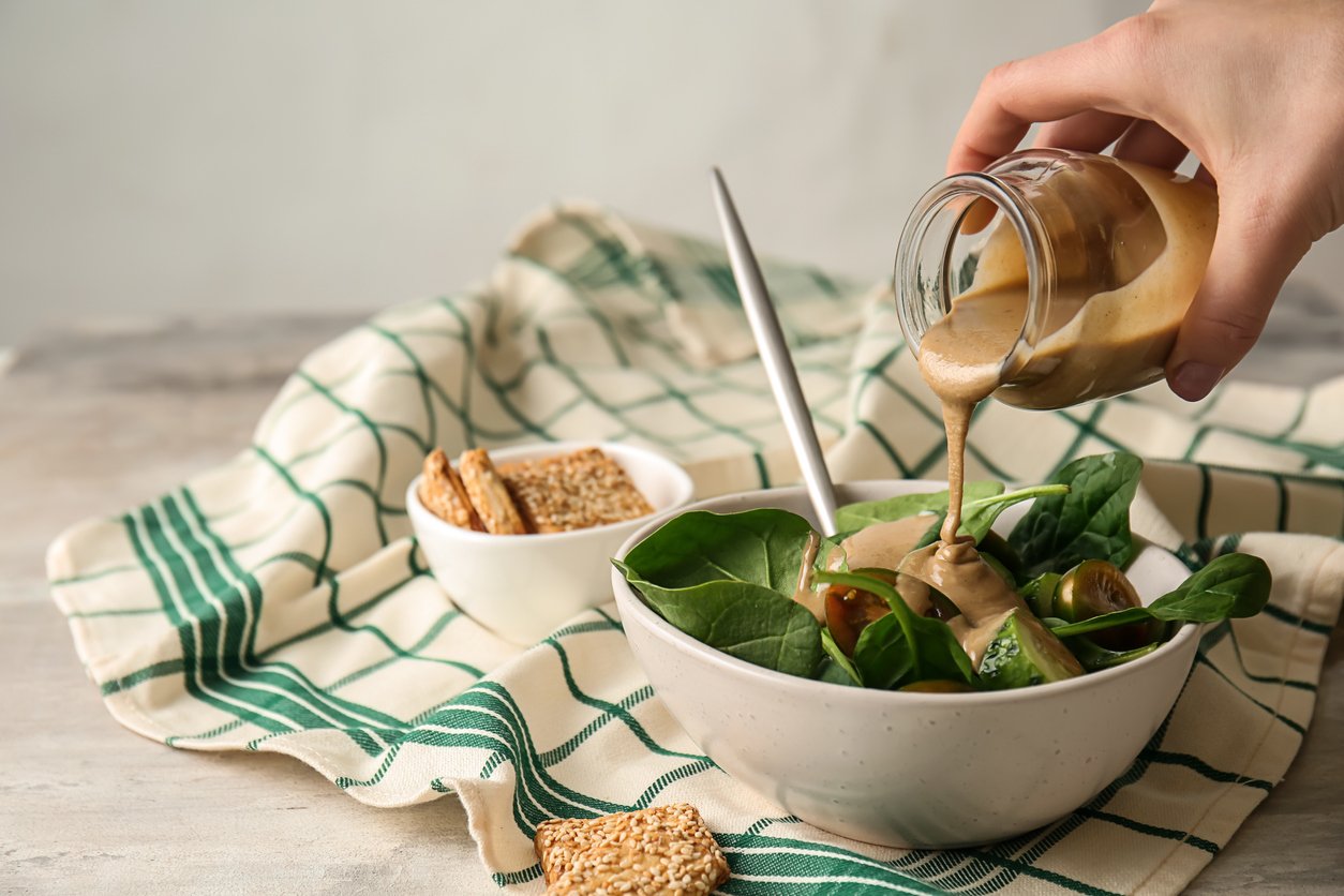 Pouring of tasty tahini from jar onto fresh vegetables in bowl