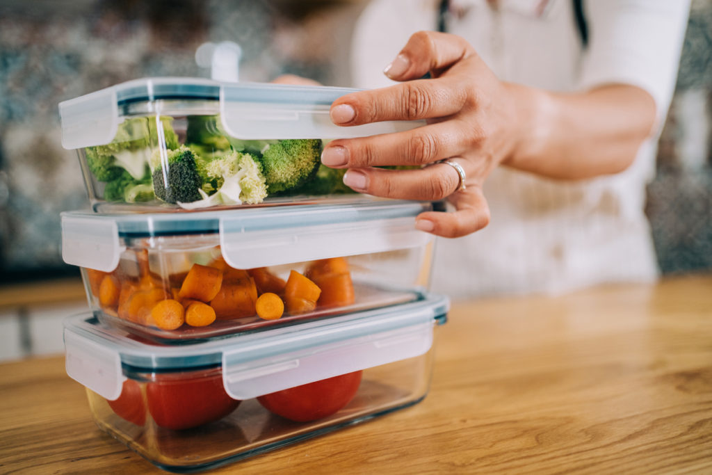 Hands holding glass airtight storage containers with fresh raw vegetables.
