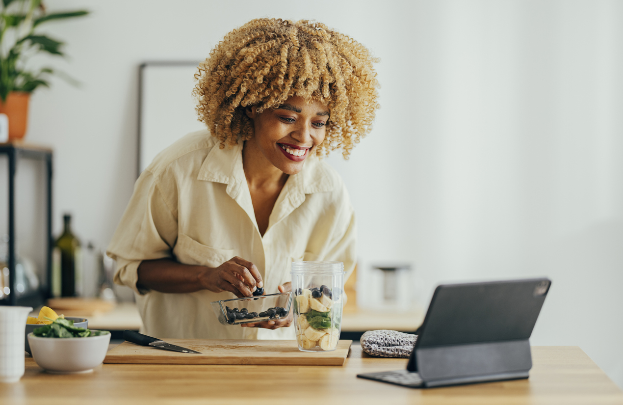 Afro-American woman with curly hair leaning over the table with a big smile on her face to see something on her tablet while cutting fruit to prepare a healthy breakfast.