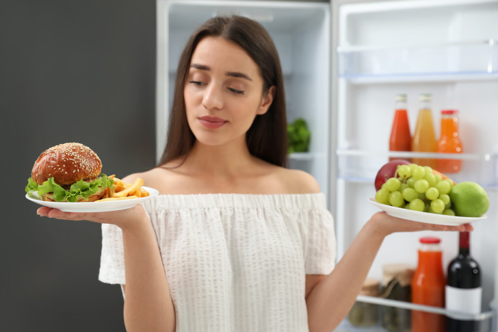 Woman choosing between fruits and burger with French fries near refrigerator in kitchen