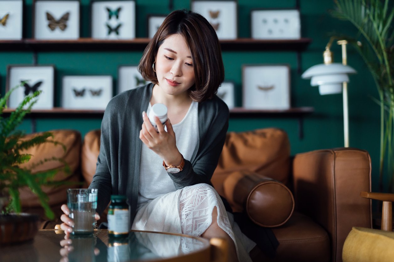 Young Asian woman taking medicines with a glass of water on the coffee table, reading the information on the label of her medication at home.