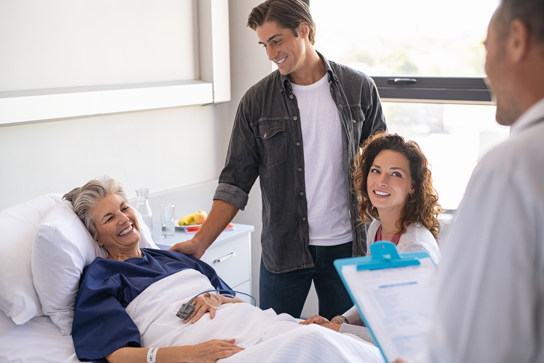 Doctor holding clipboard talking to family and smiling patient. Happy senior woman lying on hospital bed with lovely son and daughter visiting and talking to doctor. Professional friendly physician gives the results of the medical report to patient's family members.
