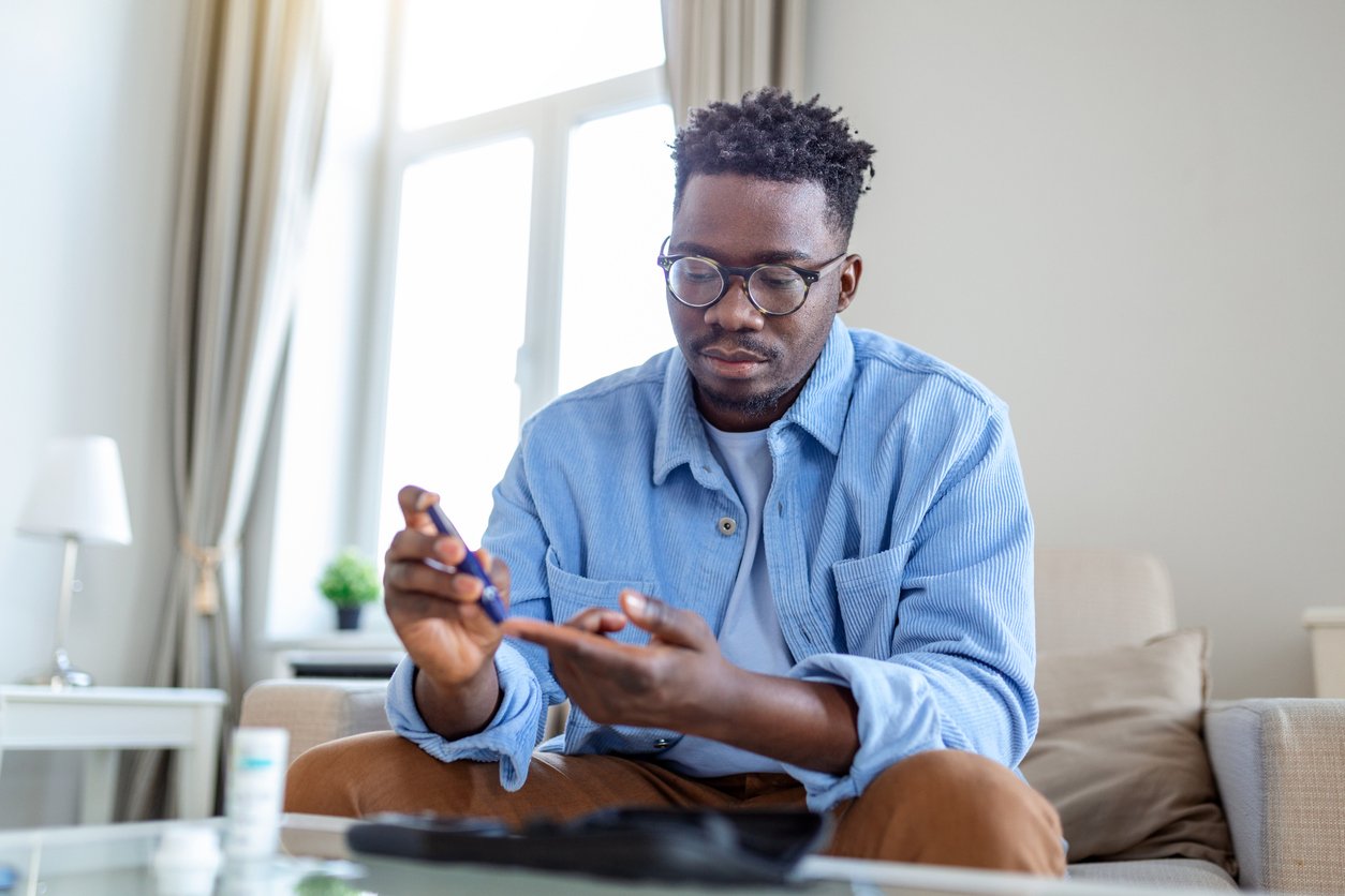 African man is sitting at the sofa and taking blood from his finger due to diabetes. The daily life of a man of African-American ethnicity person with a chronic illness who is using glucose tester.